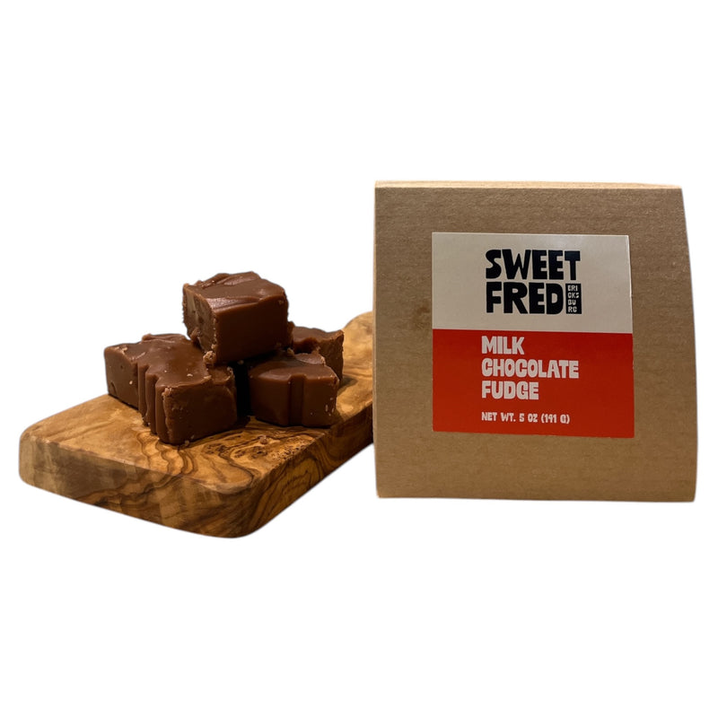 A photo of milk chocolate fudge plain 5oz package with the brand name 'SWEET FRED' visible on the packaging. The product is displayed next to a wooden board on which the fudge pieces are placed.